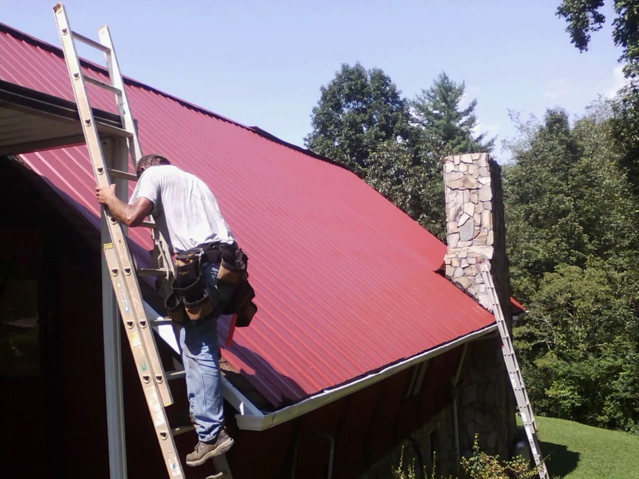 Roofer on a ladder beside a red metal roof and stone chimney — Soffit & Fascia Repair by Roofing Brothers in Tiplersville